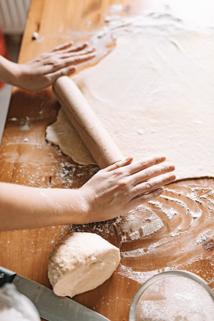 Person kneading and rolling dough with flour on a wooden kitchen table.